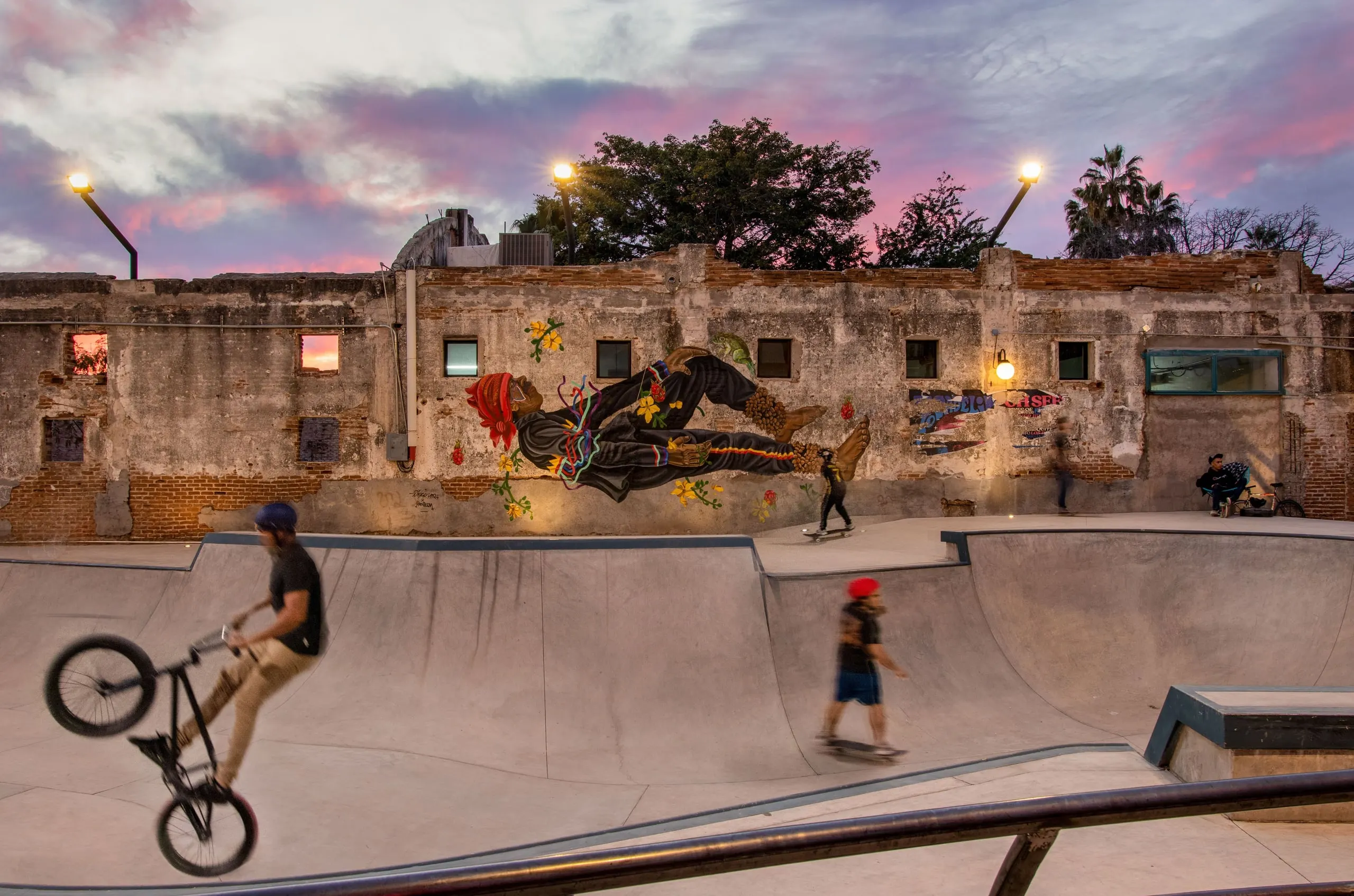 Skatepark con ciclistas y patinadores al atardecer, frente a un muro histórico con mural